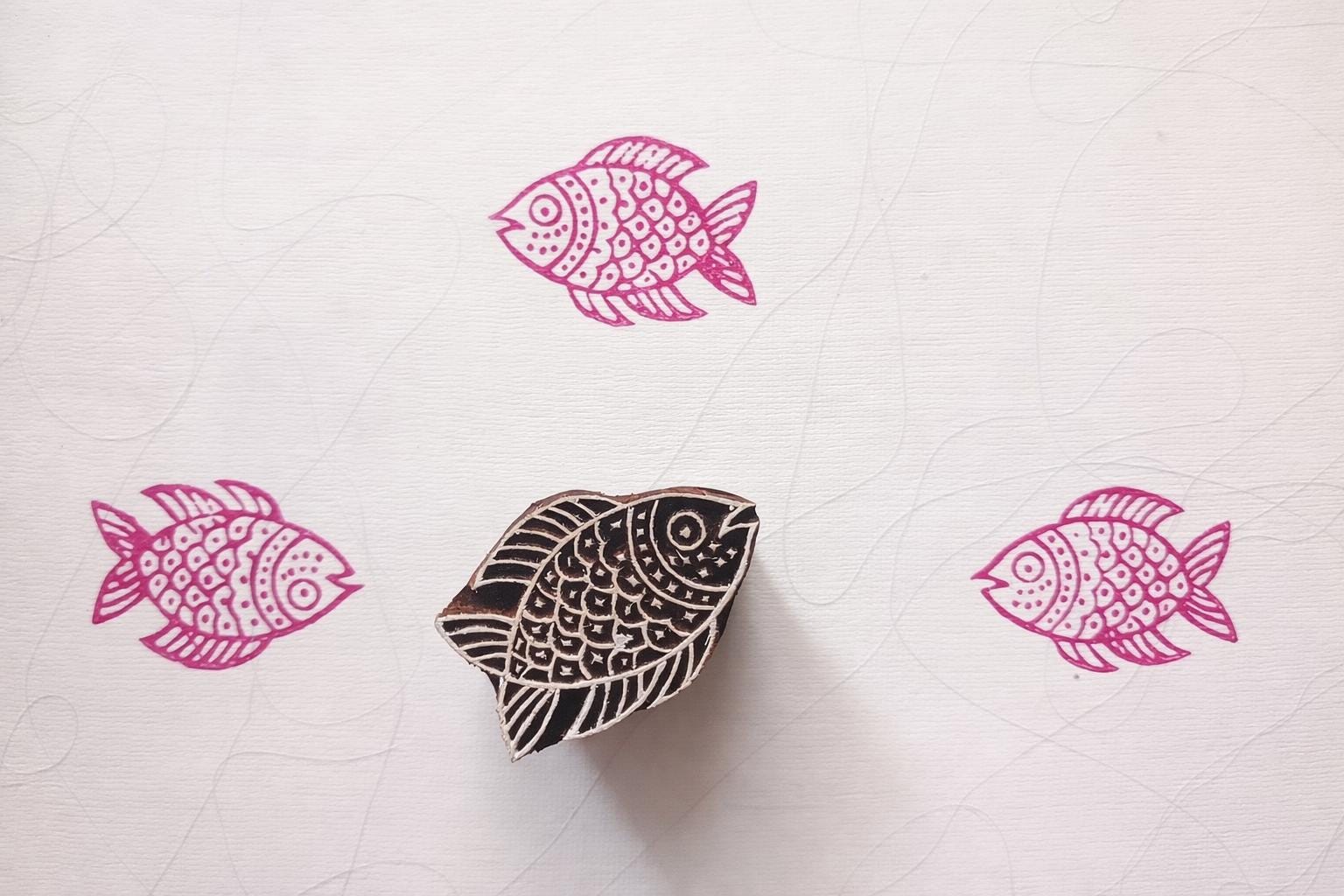 Hand carved wooden fish block printing stamp creating pink fish prints on white textured paper.