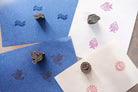 Hand carved wooden block printing stamps creating bird, fish, moon and wave prints on colored handmade paper.