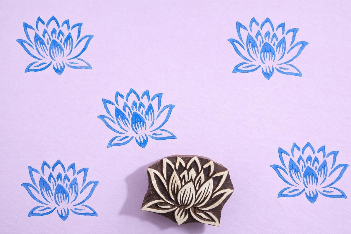 Hand carved wooden lotus flower block printing stamp creating repeating blue lotus pattern on pastel background.