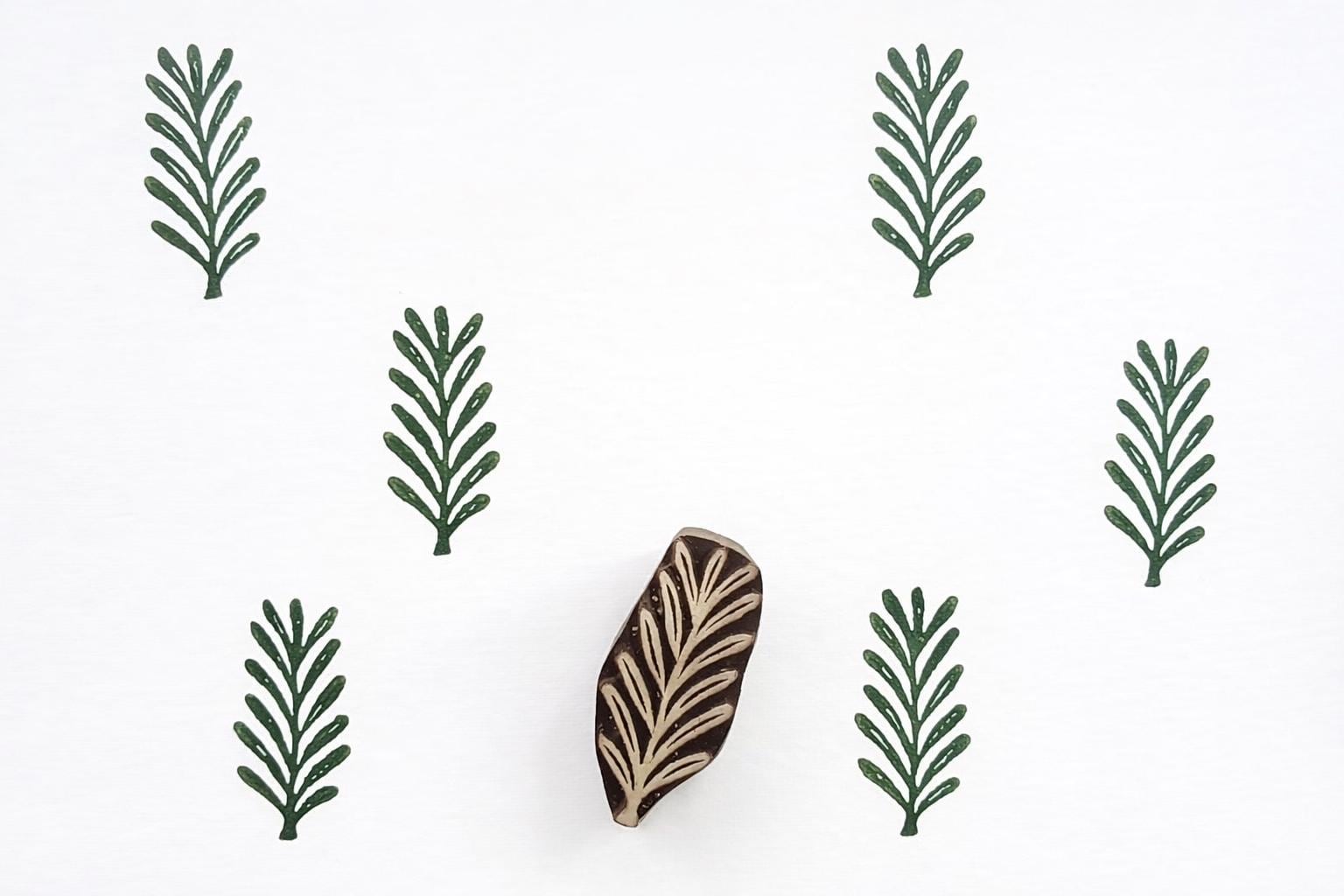 Hand carved wooden fern leaf block printing stamp creating repeating green botanical pattern on white background.