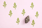 Hand carved oval wooden leaf stem block printing stamp creating light green repeating botanical pattern on soft pink background.