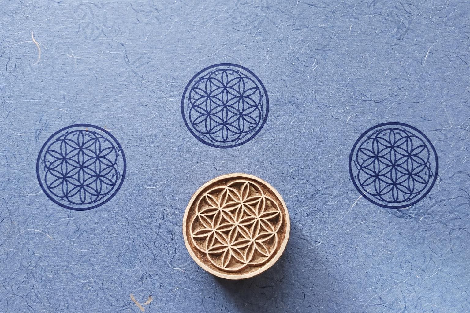 Hand carved wooden Flower of Life block printing stamp creating circular geometric prints on blue handmade paper.