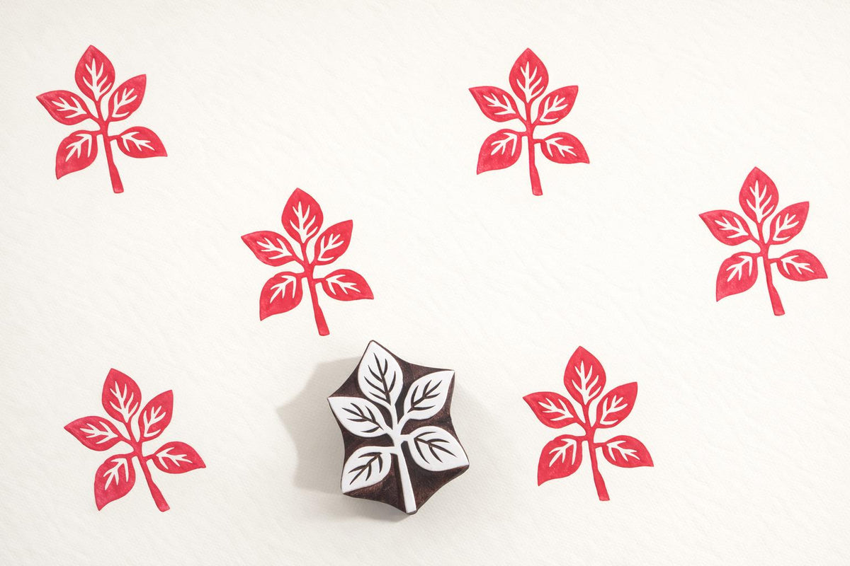 Hand carved four-leaf wooden block printing stamp creating repeating red botanical motif pattern on cream fabric.
