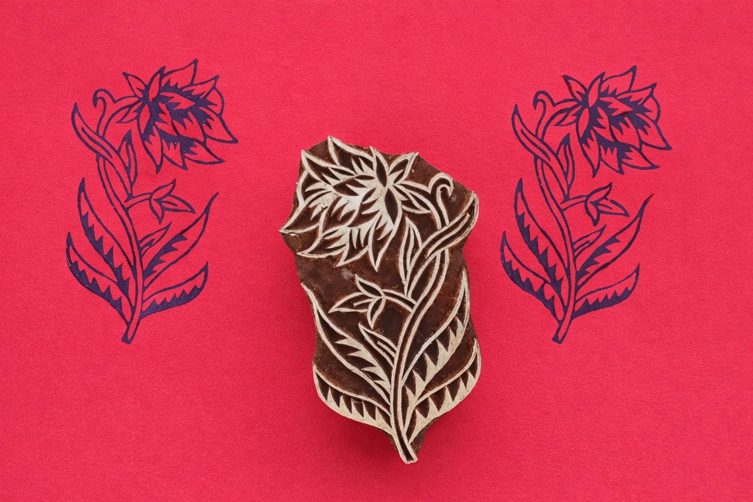 Hand carved wooden block printing stamp featuring large layered botanical flower with flowing leaves creating dark blue print on red background.