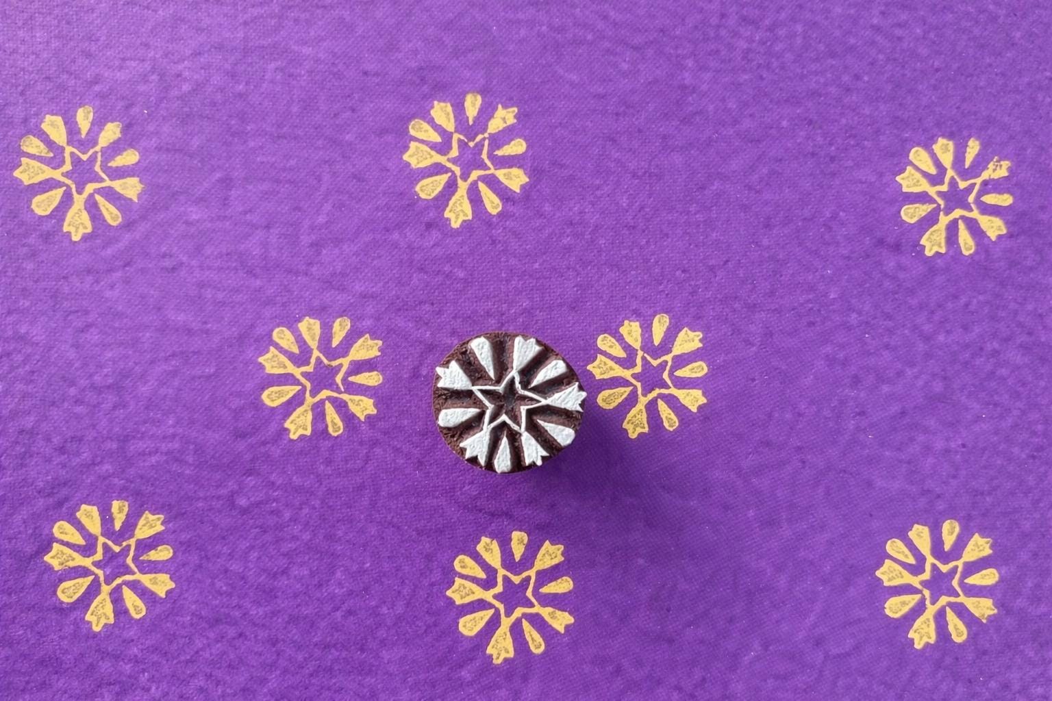 Wooden starburst flower block printing stamp making yellow prints on purple paper.