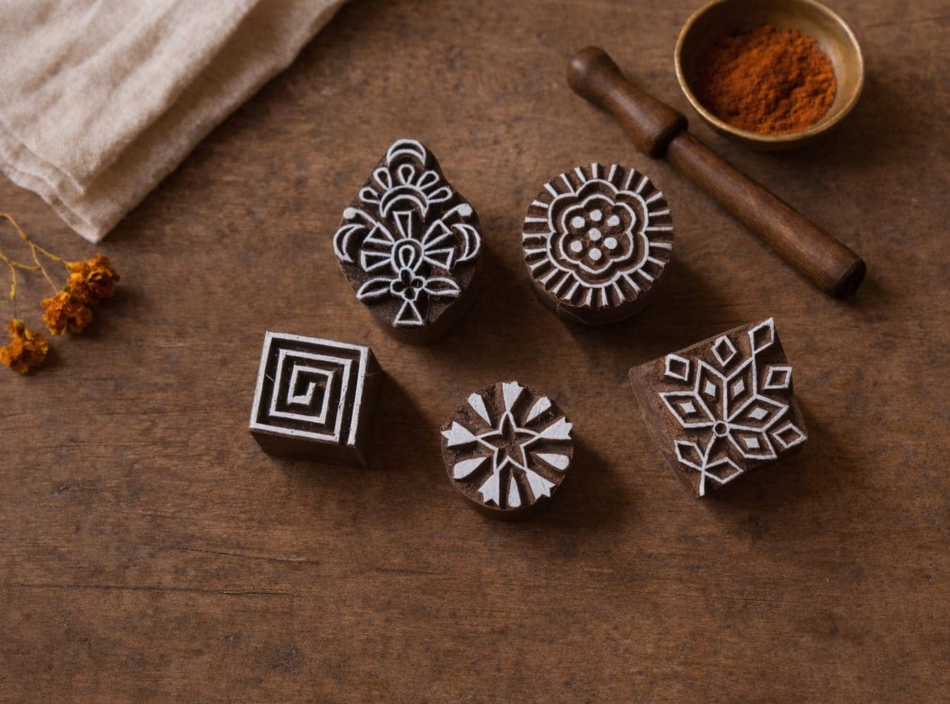 Five hand carved wooden block printing stamps on a wooden table showing Indian textile motifs including a spiral square, sunburst flower, floral rosette, geometric leaf motif, and ornamental folk design.