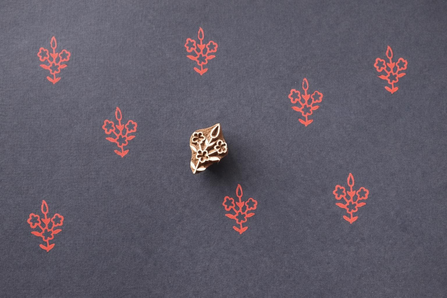 Wooden block printing stamp creating small red floral pattern prints on dark charcoal black paper.
