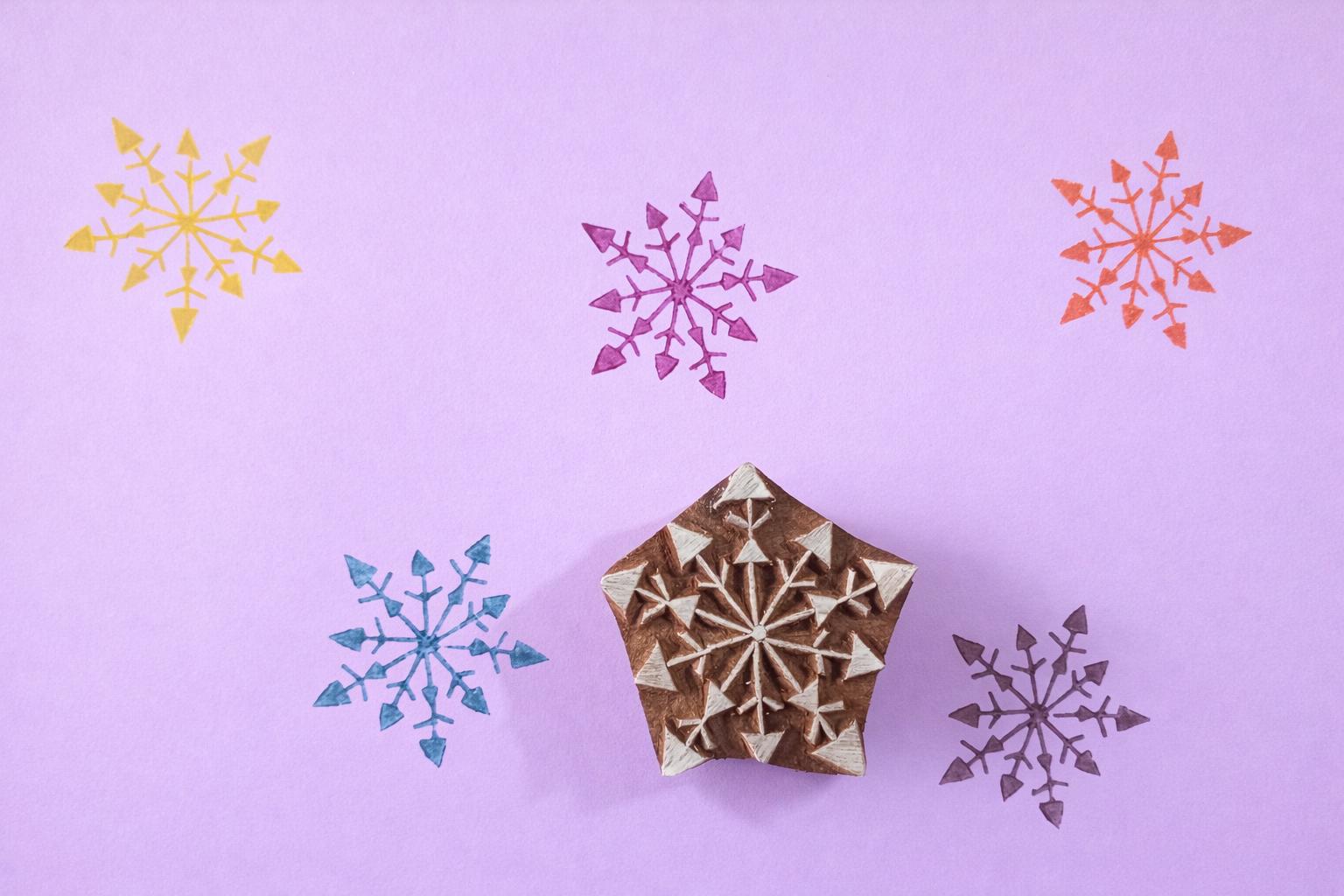 Hand carved wooden arrow snowflake block printing stamp creating colorful geometric snowflake prints on pastel background.