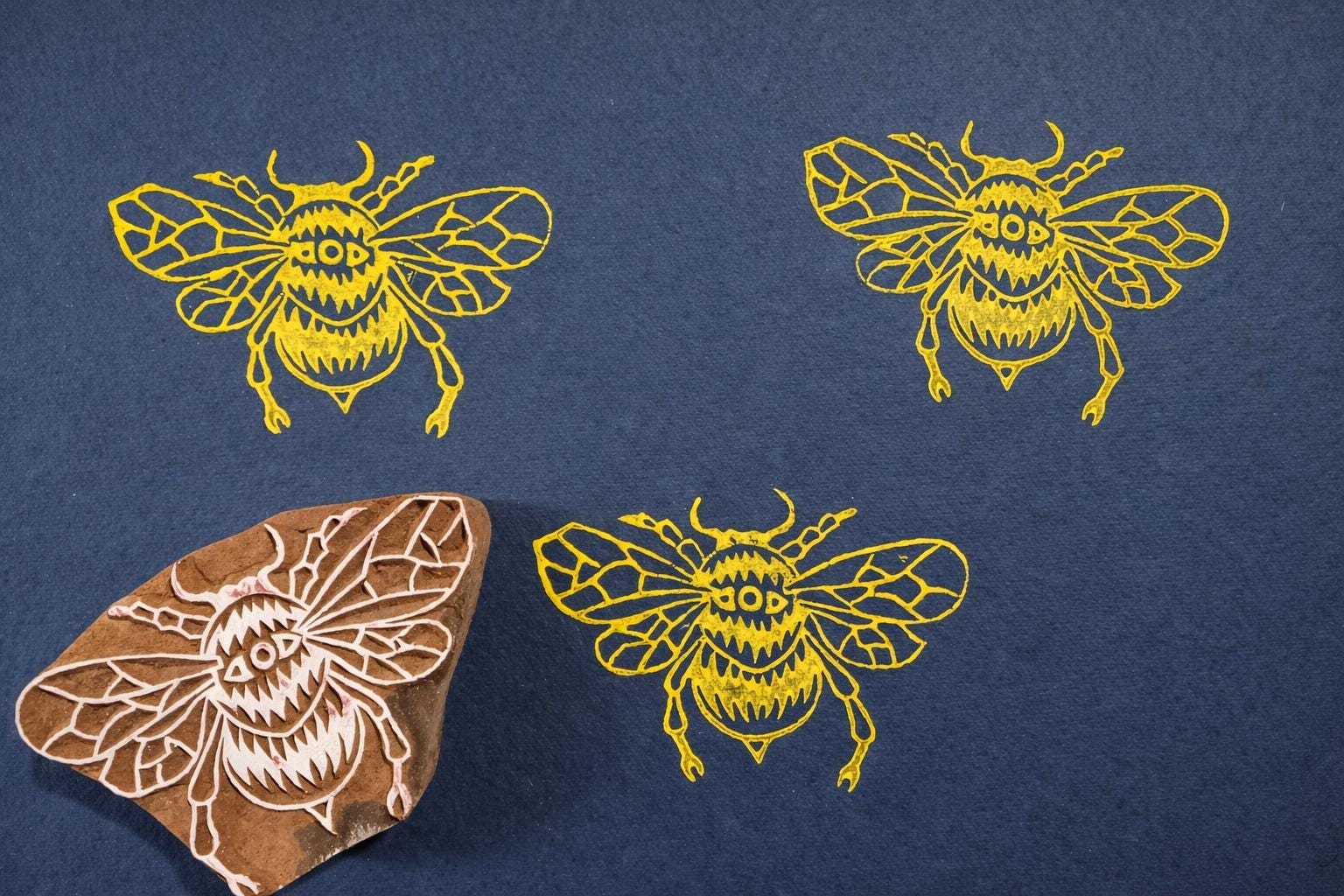 Hand carved wooden bumblebee block printing stamp creating yellow bee prints on dark blue paper.