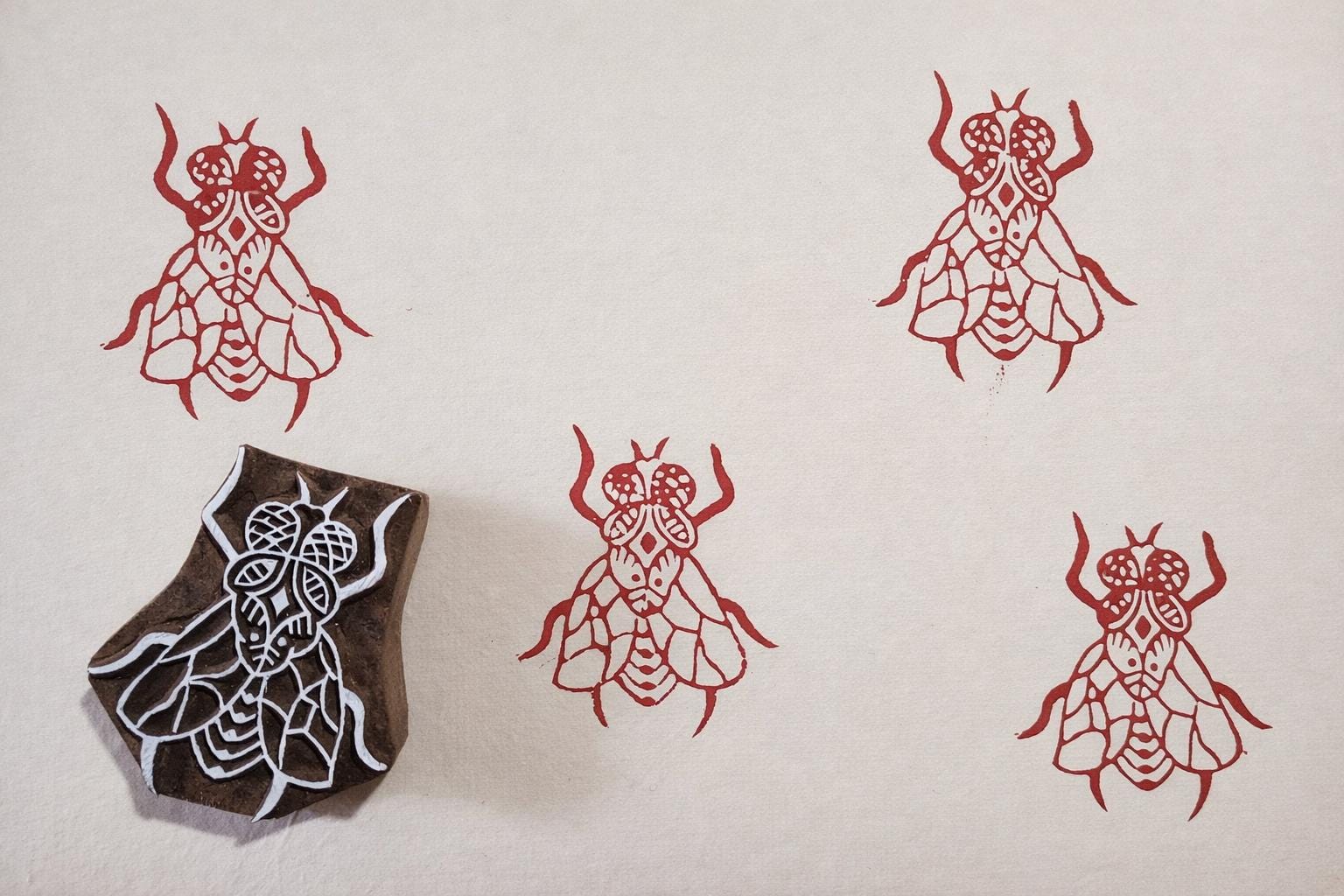 Hand carved wooden fly block printing stamp creating red insect prints on cream paper.