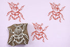 Hand carved wooden beetle block printing stamp creating coral insect prints on pastel purple paper.