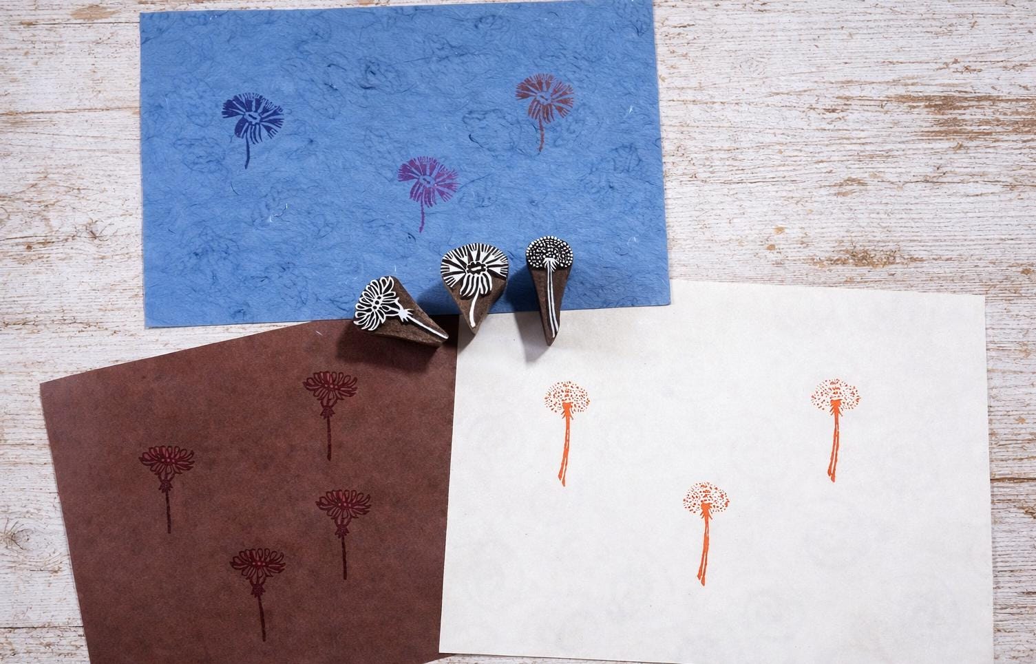 Three hand carved wooden wildflower block printing stamps displayed with printed botanical patterns on blue, brown and white paper.