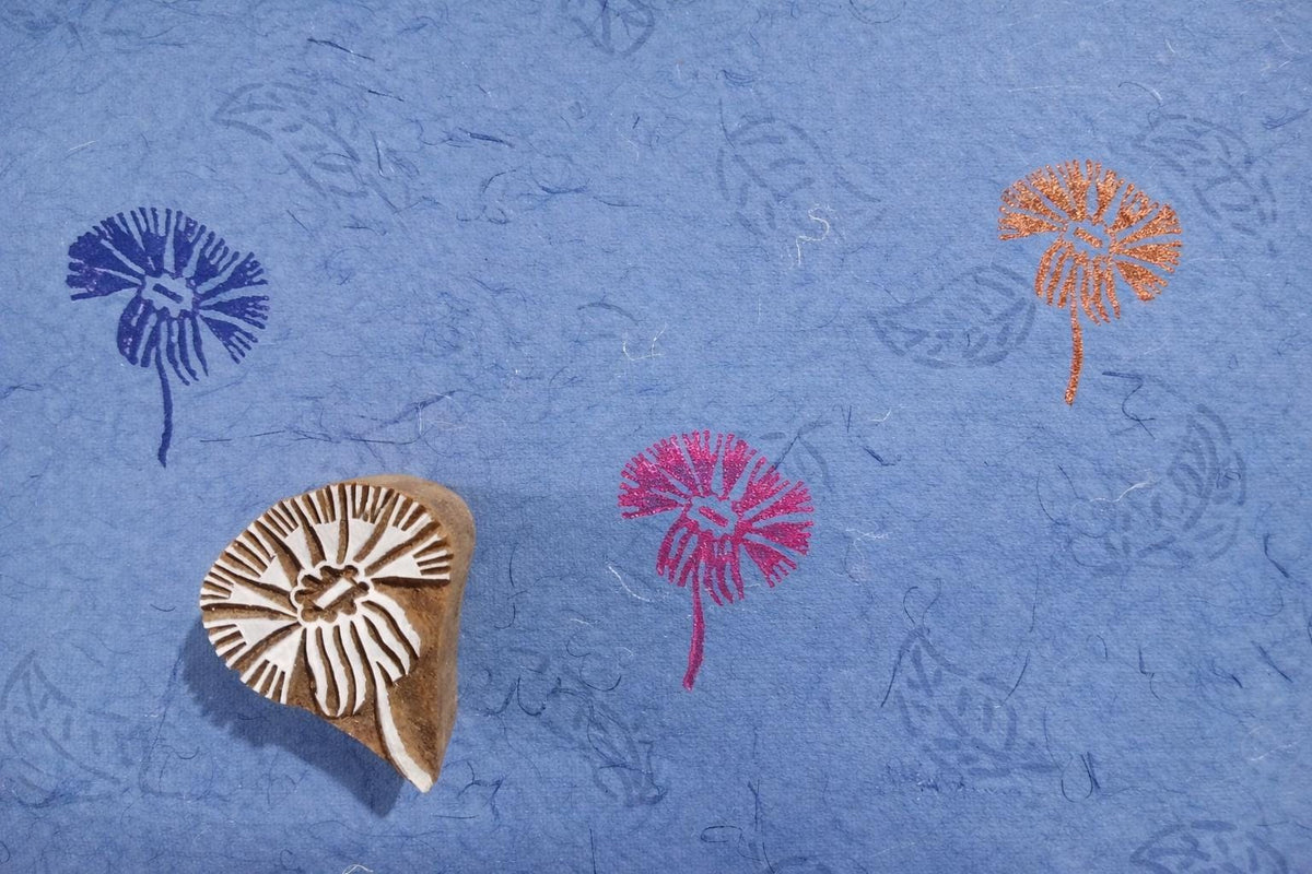 Hand carved wooden daisy bloom block printing stamp creating colorful floral prints on handmade blue paper.