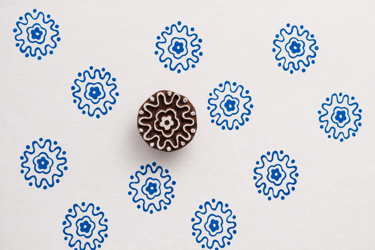 Round sunburst rosette block printing stamp creating blue floral prints on white paper background.