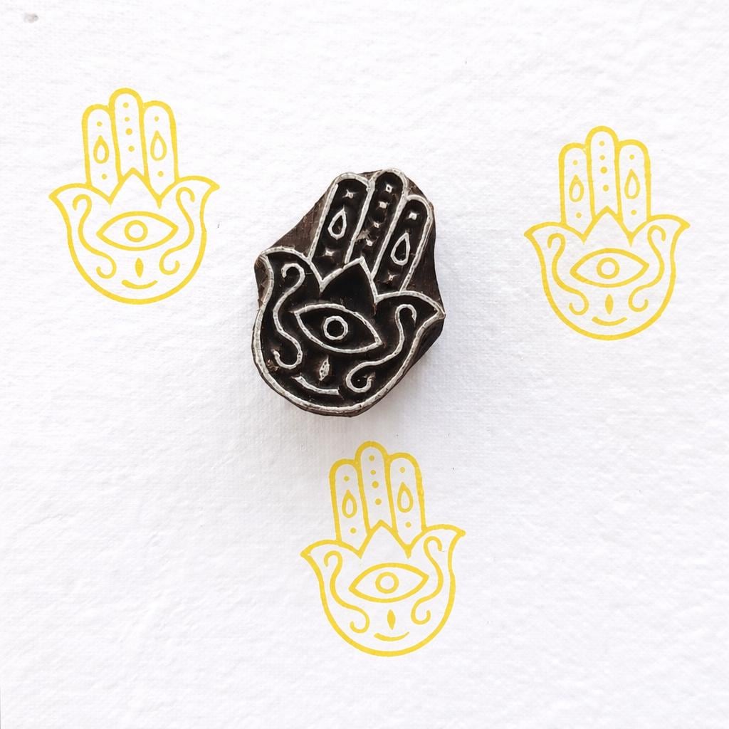 Hamsa hand wooden block printing stamp creating yellow hamsa prints on white fabric paper background.