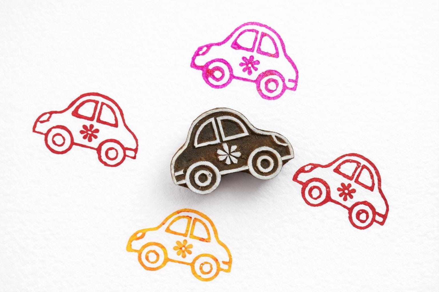 Hand carved wooden car block printing stamp creating colorful car prints in pink, red and orange on white paper.