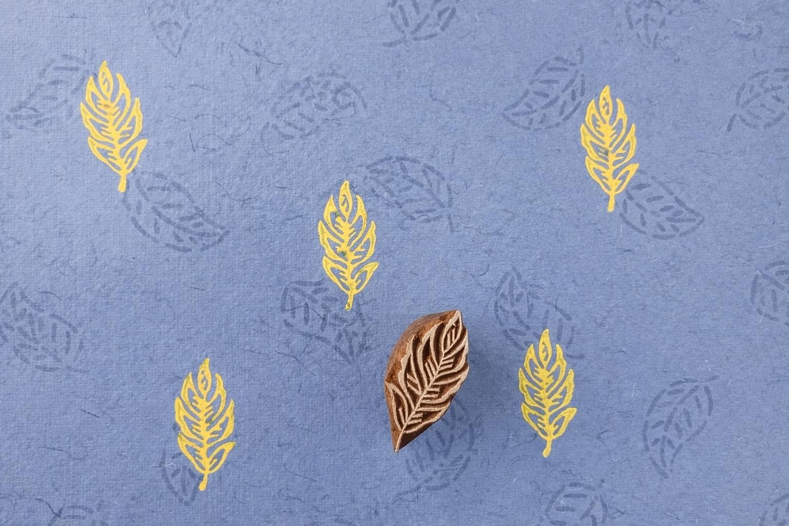 Hand carved leaf wooden block printing stamp creating yellow leaf prints on textured blue handmade paper.