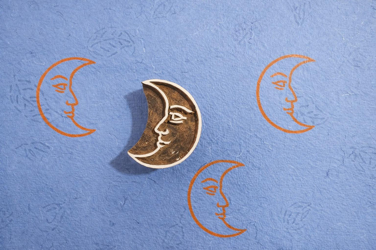 Hand carved wooden crescent moon face block printing stamp creating orange moon prints on blue handmade paper.