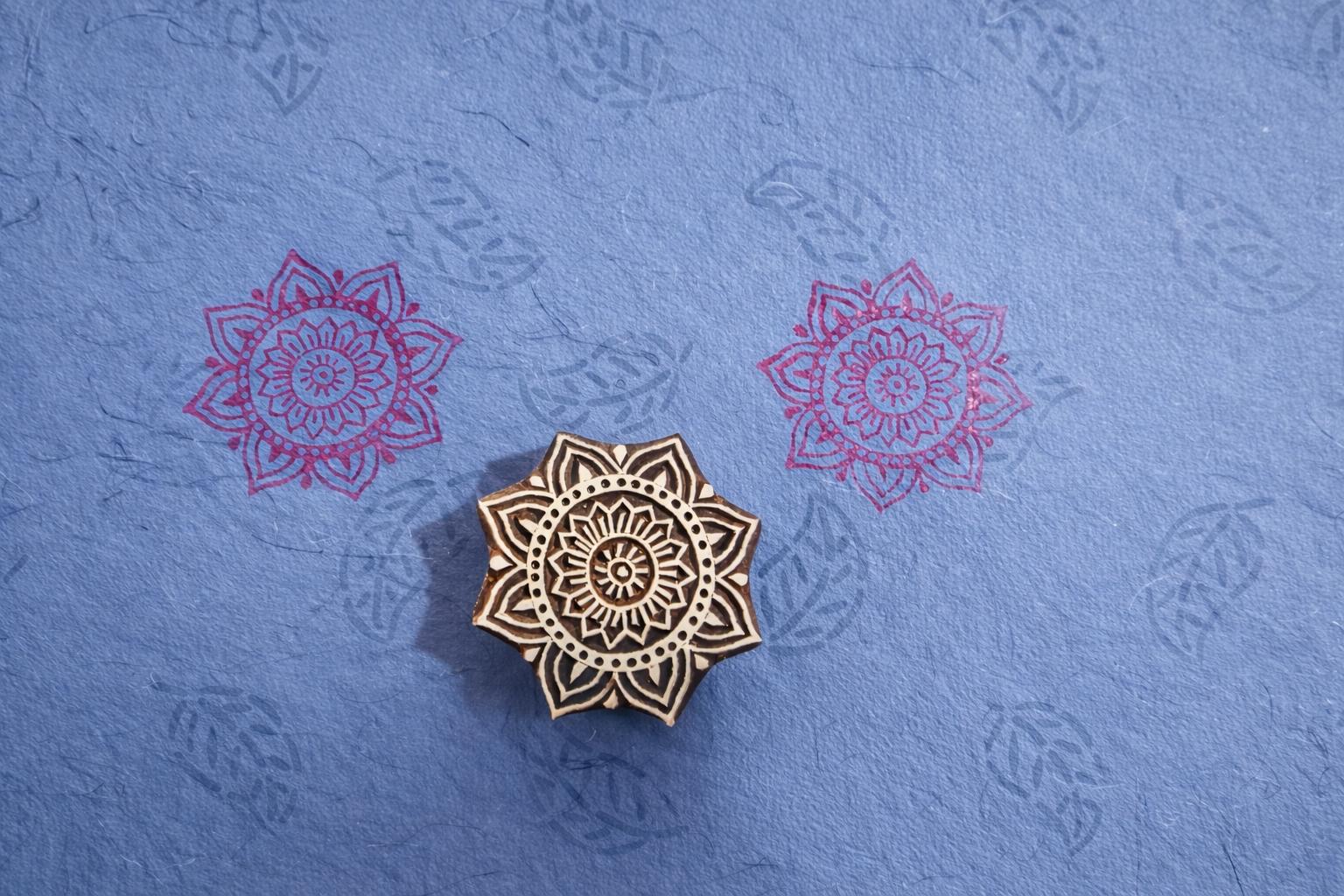 Hand carved wooden mandala flower block printing stamp creating pink mandala prints on blue handmade paper.