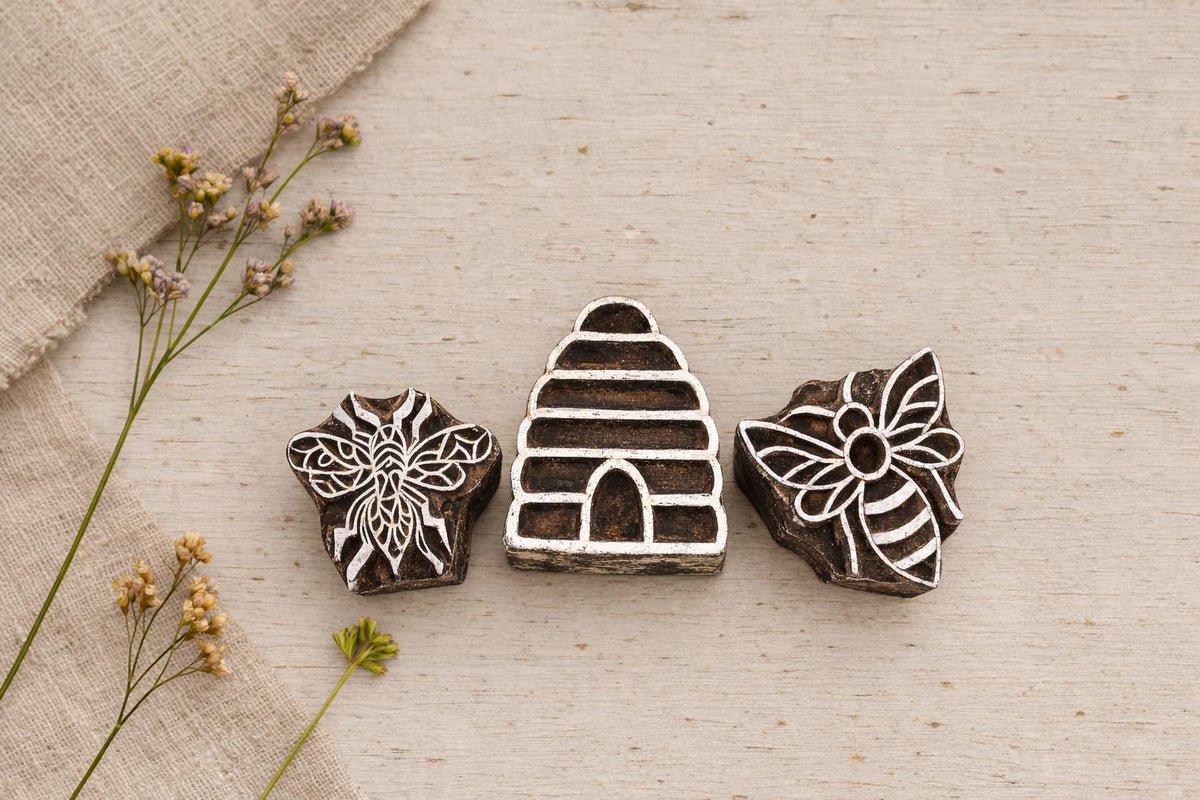 Hand carved wooden block printing stamp set featuring honeybee, beehive and floral bee motifs displayed on rustic table with linen cloth and dried flowers.