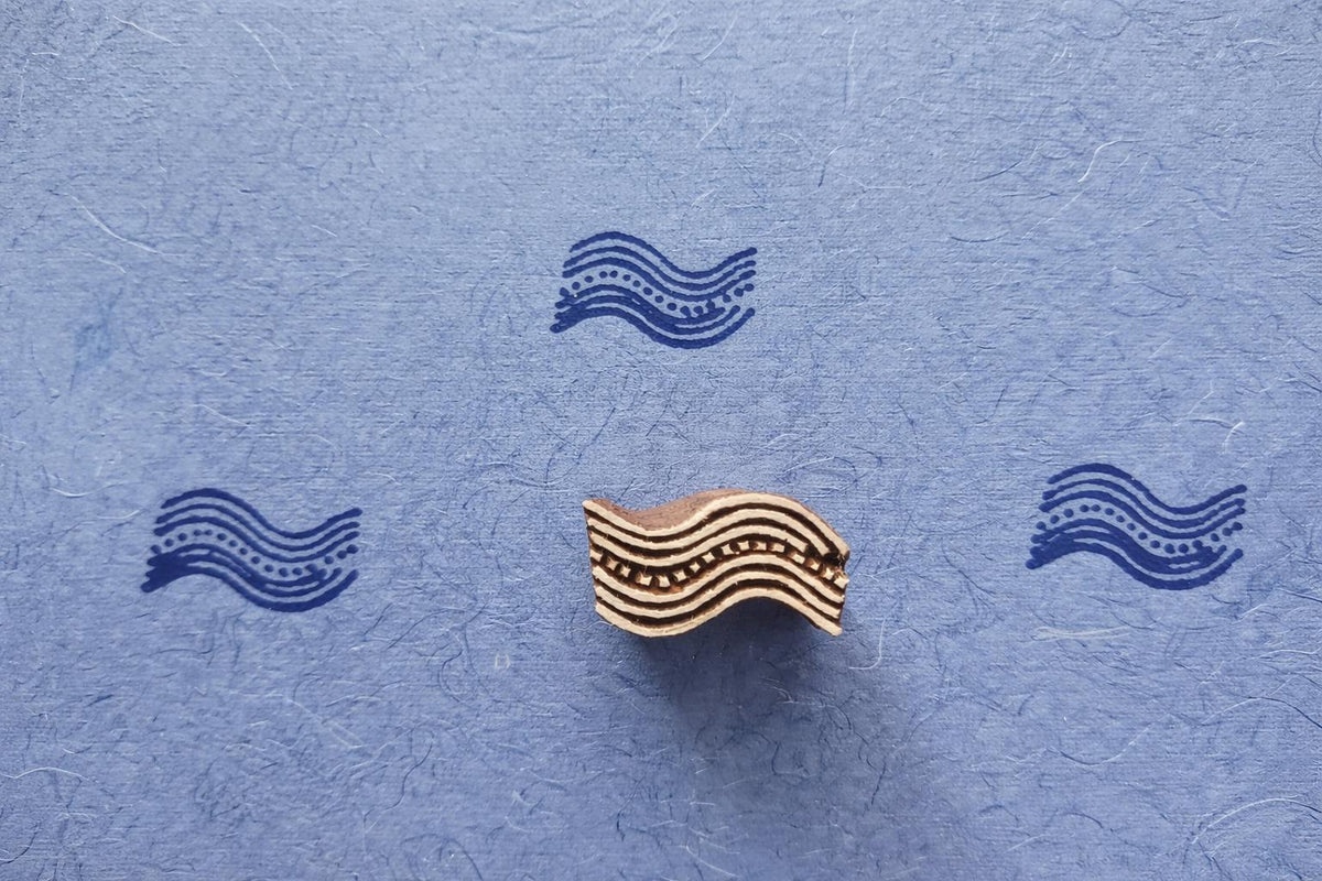Hand carved wooden wave pattern block printing stamp creating blue wave prints on textured paper.