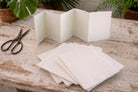 Handmade off-white deckle edge paper folded into an accordion strip on a rustic wooden table, showing soft cream paper color and textured torn edges