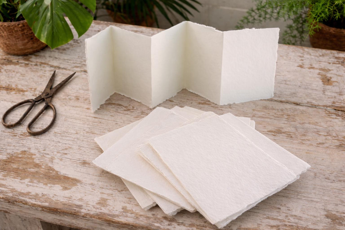 Handmade off-white deckle edge paper folded into an accordion strip on a rustic wooden table, showing soft cream paper color and textured torn edges
