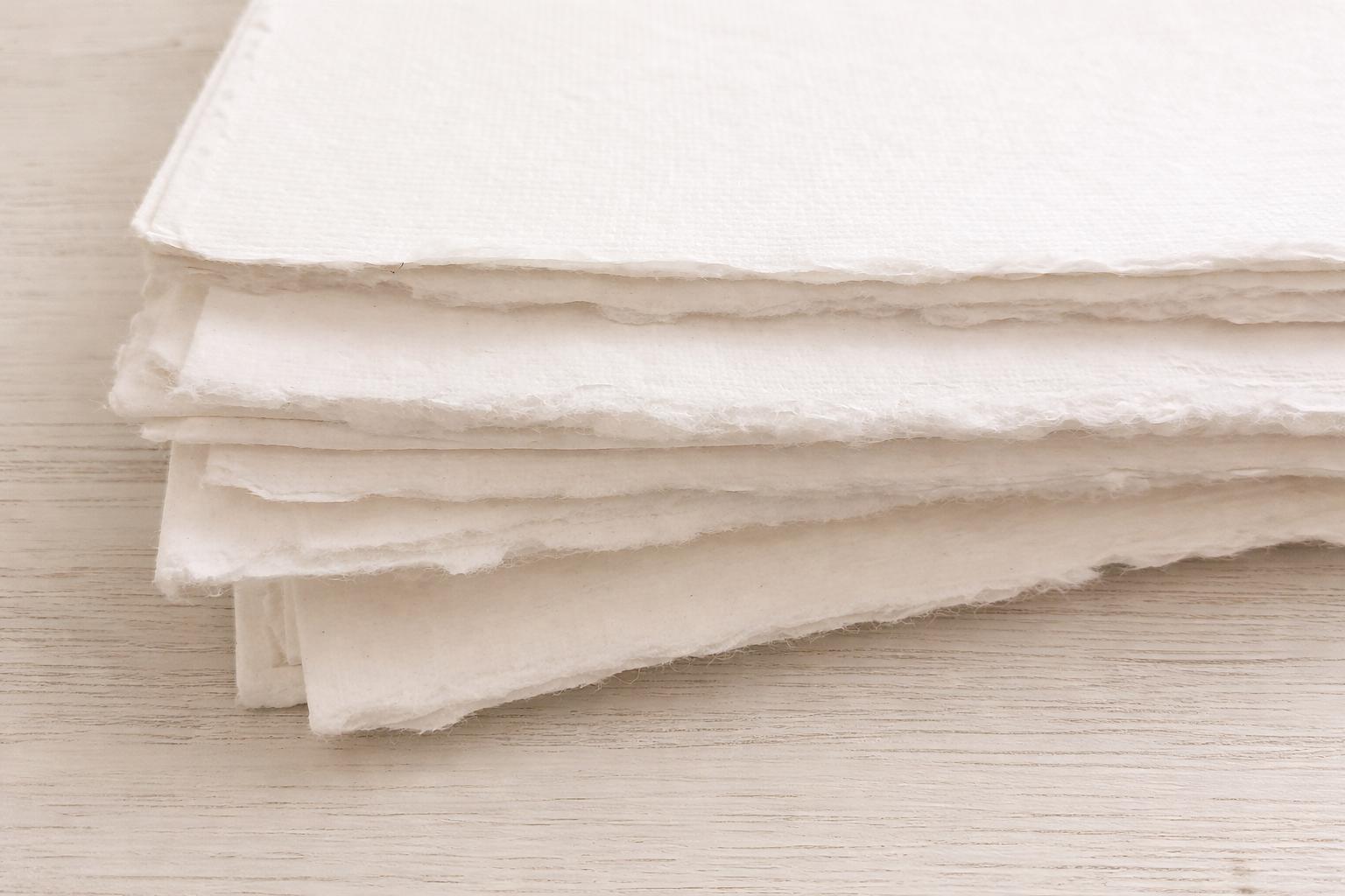 Stack of handmade off-white deckle edge paper sheets viewed from the side, highlighting rough torn edges and thick textured cream paper