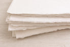 Stack of handmade off-white deckle edge paper sheets viewed from the side, highlighting rough torn edges and thick textured cream paper