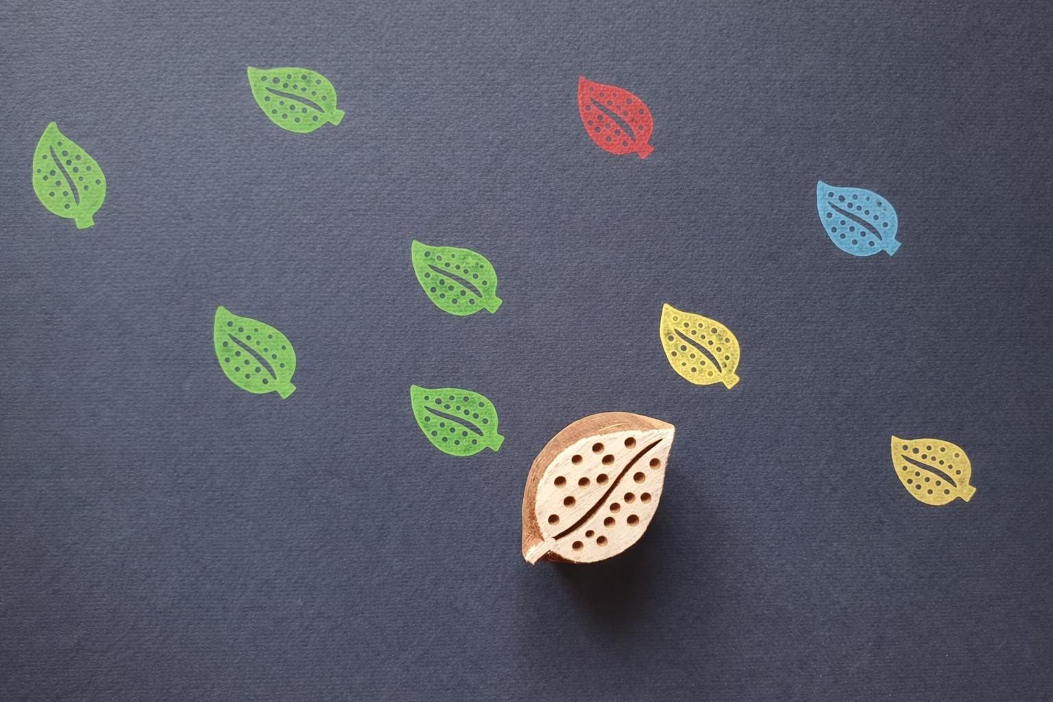 Leaf block printing stamp creating colorful repeated leaf patterns on dark background paper.
