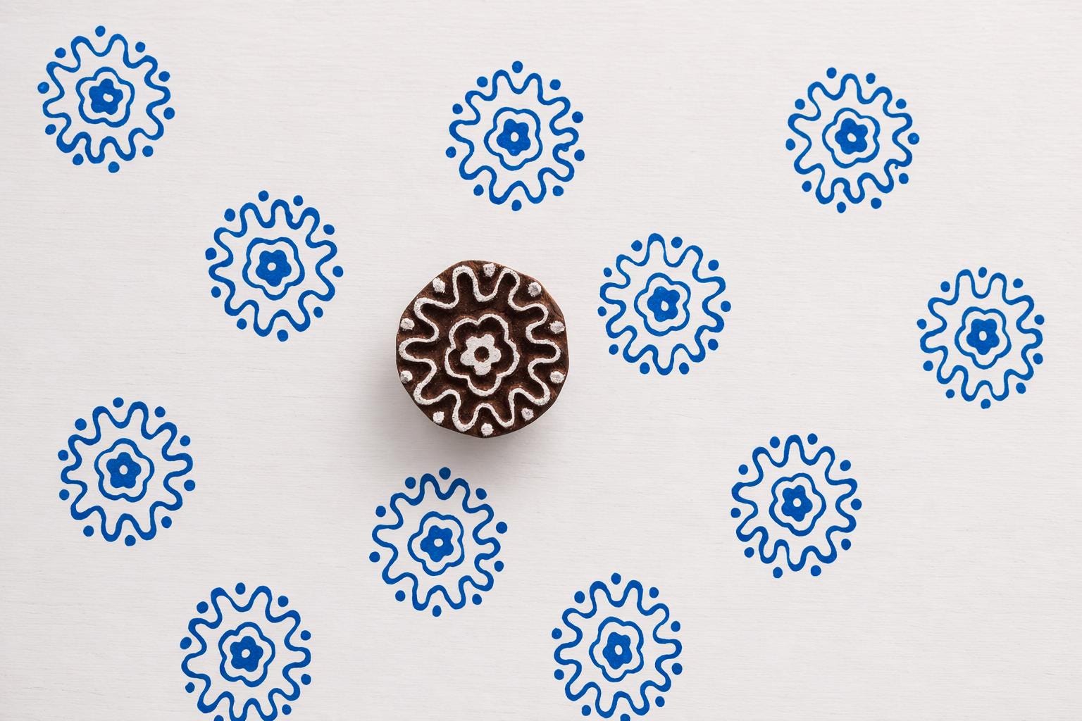 Round sunburst rosette block printing stamp creating blue floral prints on white paper background.