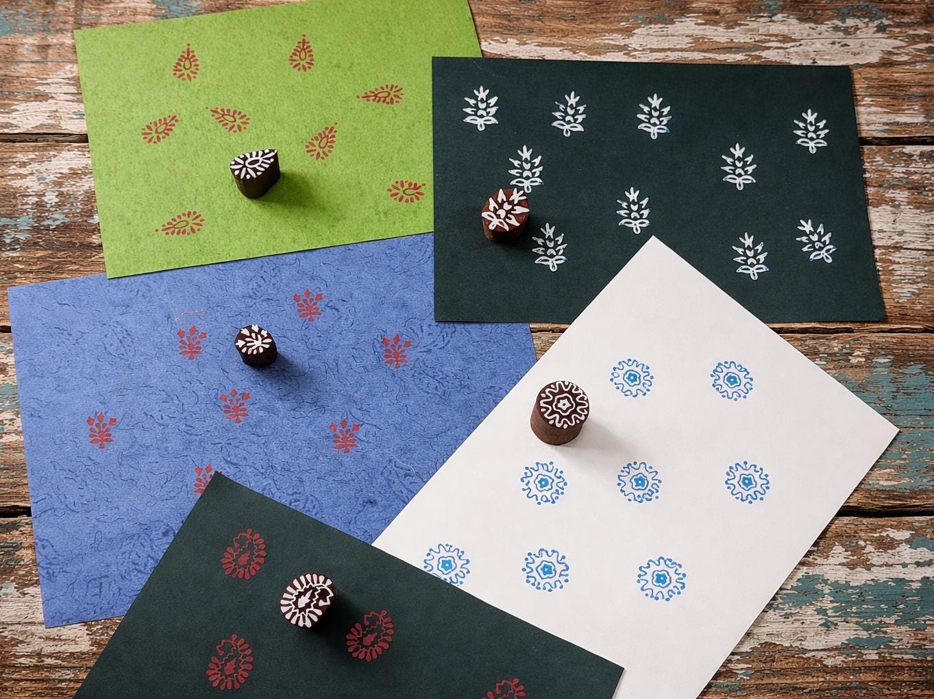 Hand carved Indian wooden block printing stamp set displayed on colorful handmade paper sheets showing red, blue and white printed patterns on green, blue and white paper.