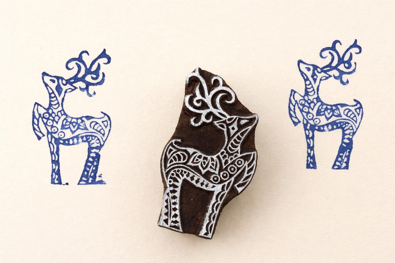 Hand-carved wooden deer stamp with intricate folk art design, shown stamping blue deer patterns on a warm beige background.