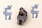 Hand-carved wooden deer stamp with intricate folk art design, shown stamping blue deer patterns on a warm beige background.