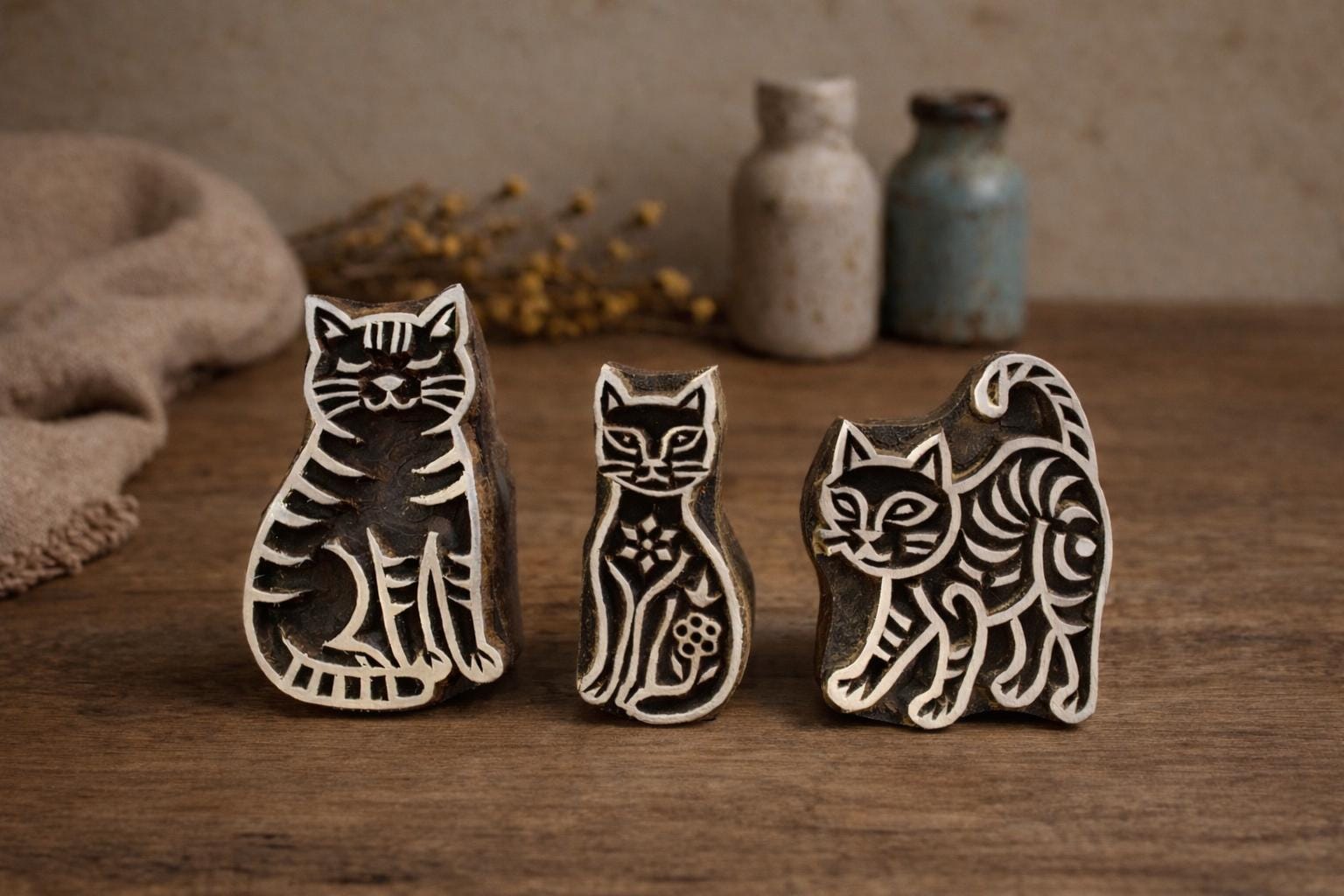 Hand carved wooden cat block printing stamps set featuring three cat designs displayed on rustic wooden table with fabric and ceramic bottles.