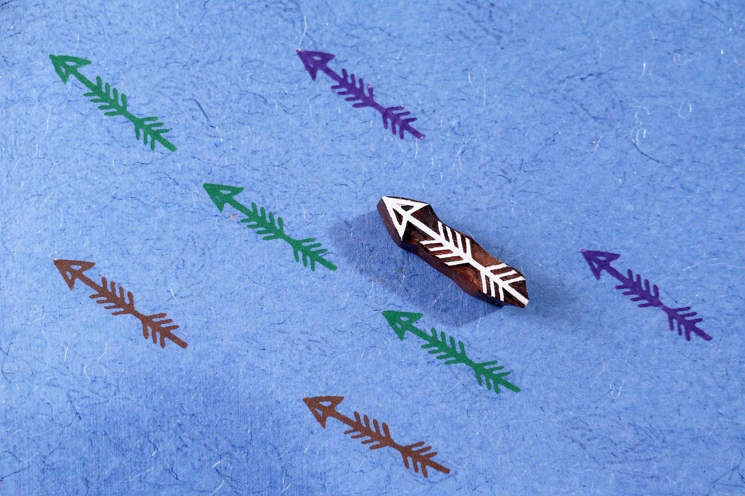 Hand carved arrow stamp used to print multicolor arrow patterns on textured blue paper.
