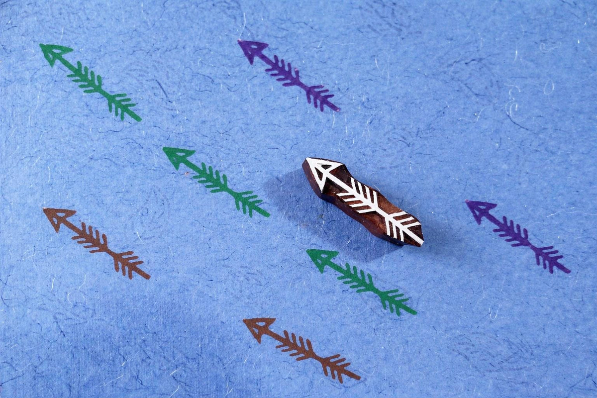 Hand carved arrow stamp used to print multicolor arrow patterns on textured blue paper.
