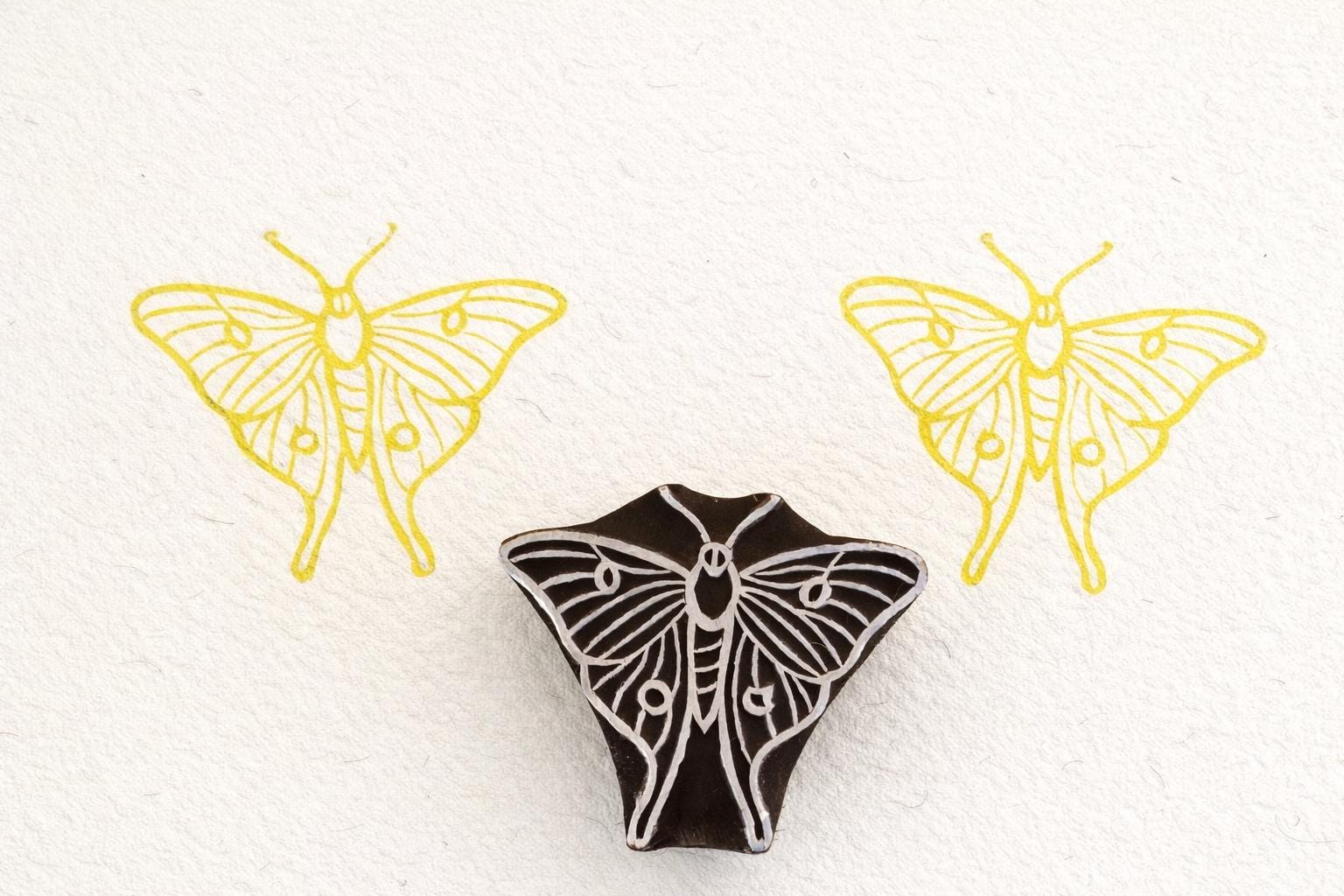Hand-carved luna moth stamp printing simple symmetrical moth pattern on paper.