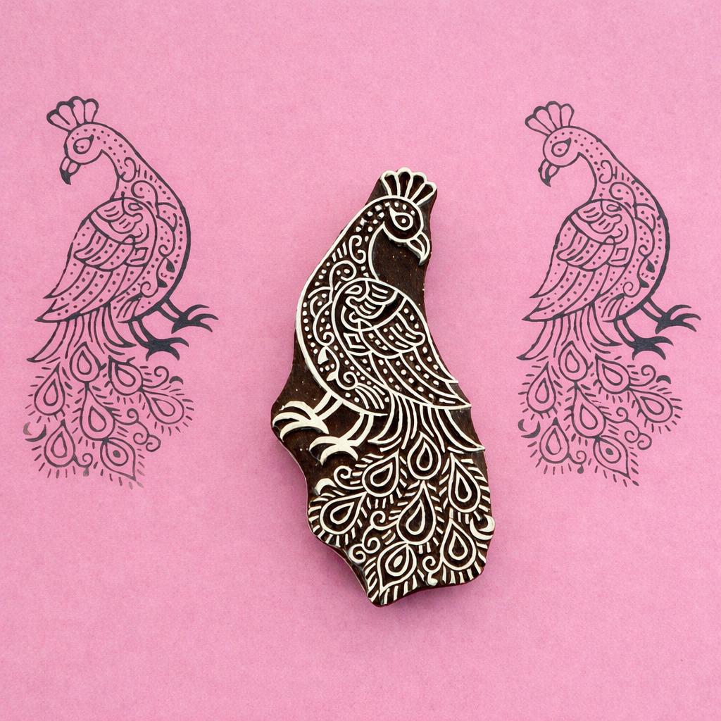 Hand carved wooden block stamp of a detailed peacock with feather pattern printed in black on pink paper