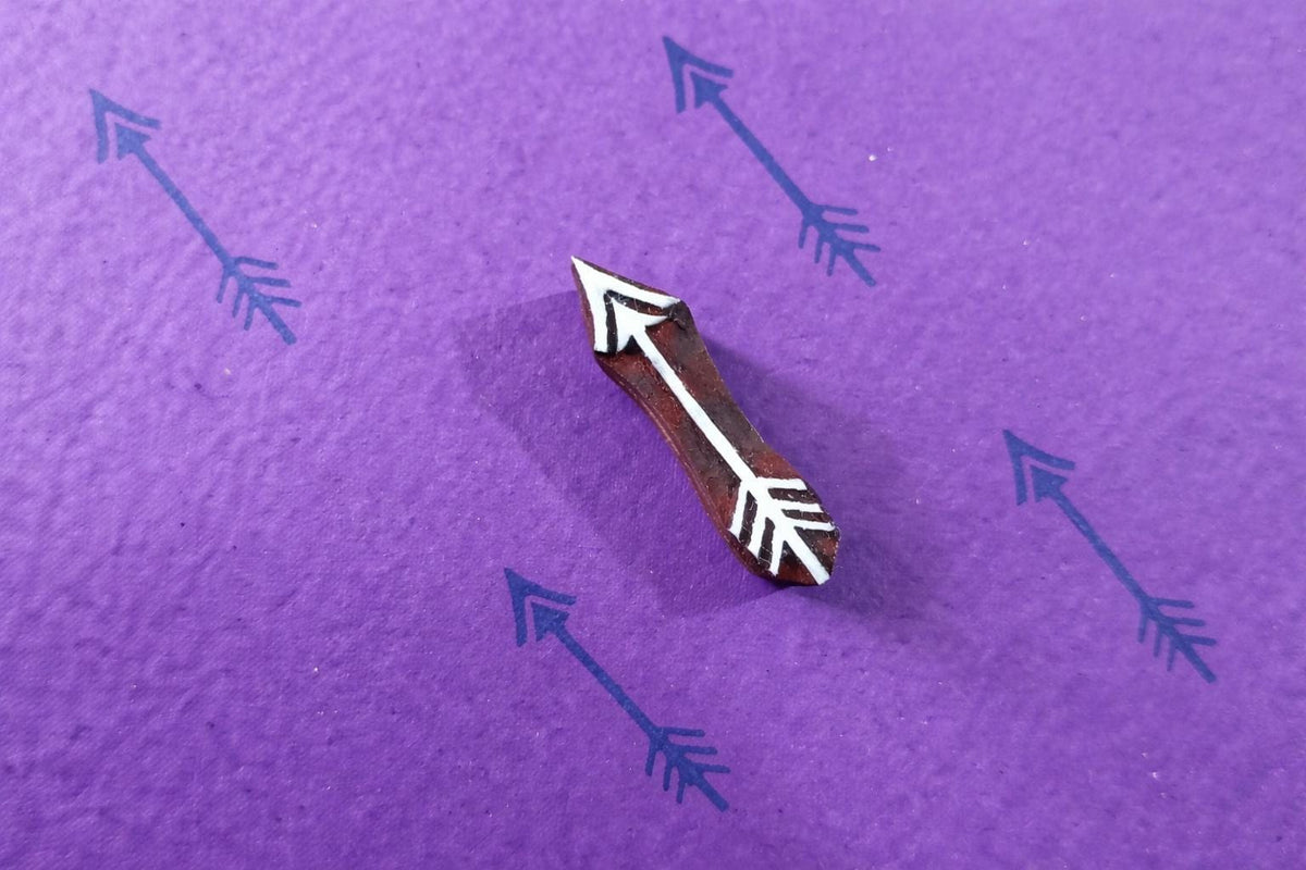 Single wooden arrow stamp creating a repeating minimalist arrow pattern on purple paper.