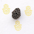 Hamsa hand wooden block printing stamp creating yellow hamsa prints on white fabric paper background.