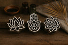 Hand carved wooden block printing stamp set featuring lotus flower, hamsa hand and mandala symbols displayed on rustic wooden table with pottery jars and dried palm leaf.