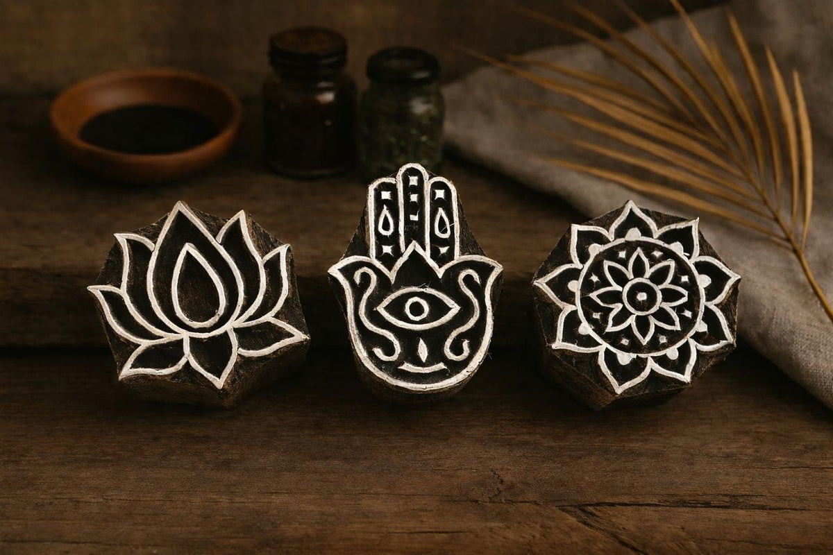 Hand carved wooden block printing stamp set featuring lotus flower, hamsa hand and mandala symbols displayed on rustic wooden table with pottery jars and dried palm leaf.