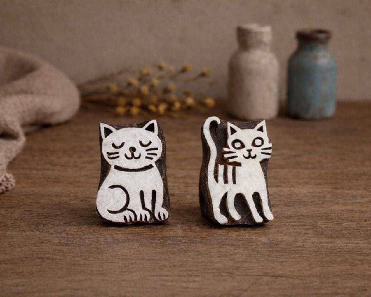 Hand carved wooden block printing stamps featuring two cat designs, sleepy curled cat and upright tail cat, displayed on rustic wooden table with fabric and ceramic bottles.