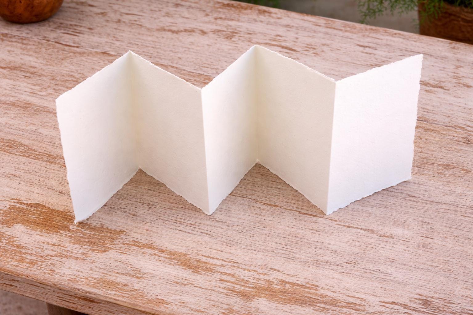 Accordion folded handmade paper in off-white cream color showing multiple panels and soft deckle edges on a wooden surface