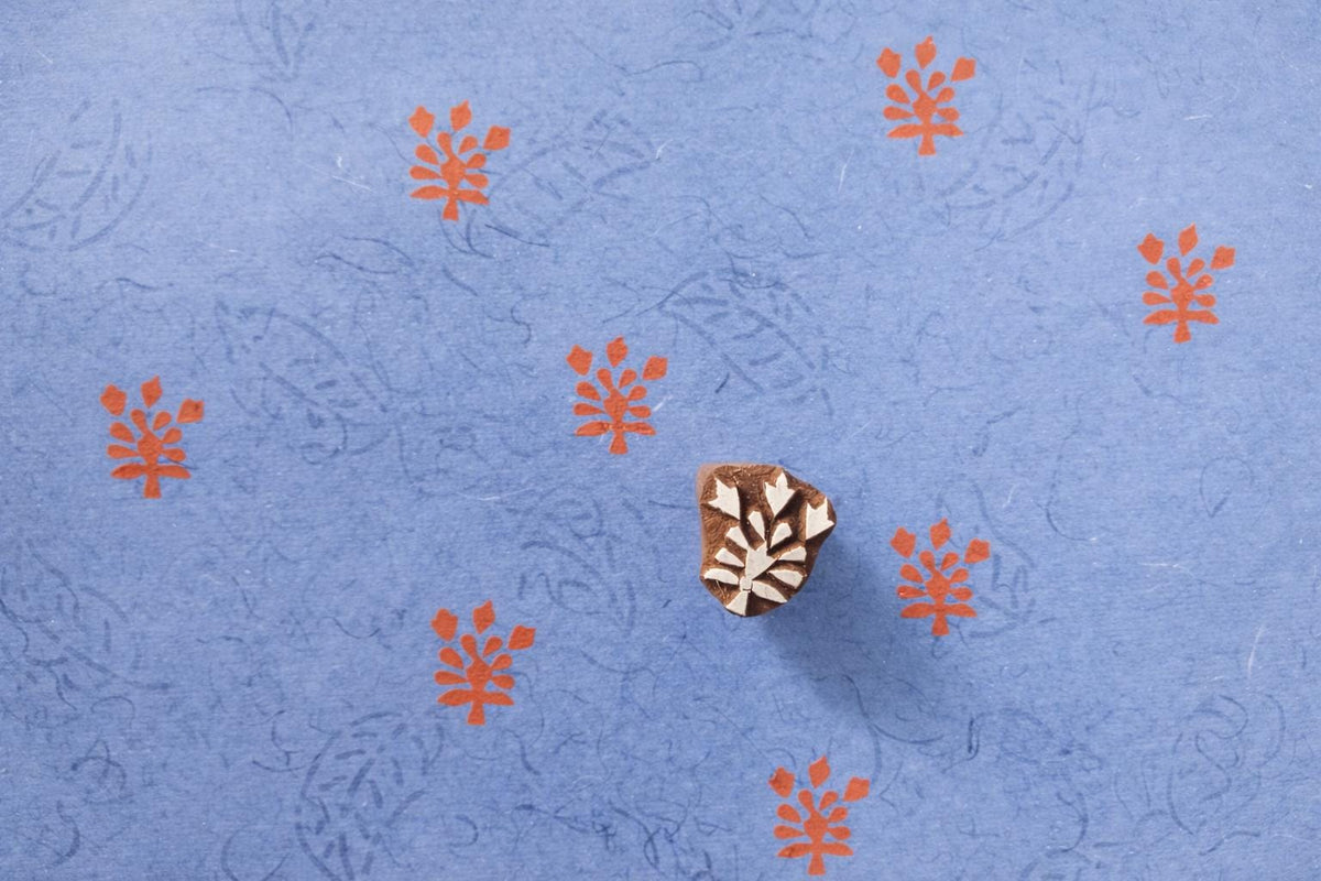 Fern branch wooden block printing stamp making orange prints on textured blue handmade paper.