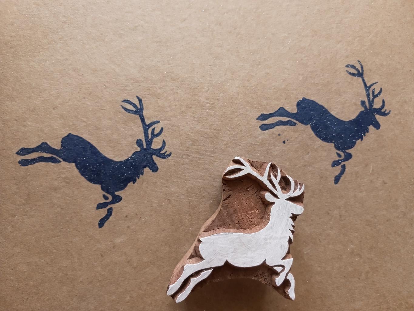 Hand-carved wooden deer block stamp with dark blue leaping stag prints on paper in a rustic woodland style