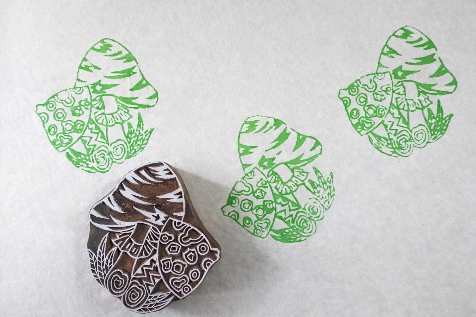 Hand-carved wooden mushroom stamp with detailed woodland design, printing repeating mushroom patterns on white paper.