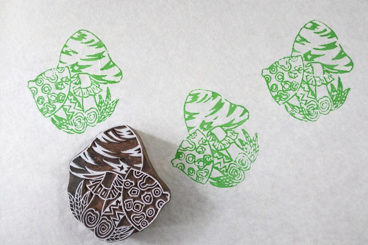 Hand-carved wooden mushroom stamp with detailed woodland design, printing repeating mushroom patterns on white paper.