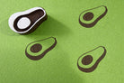 Hand-carved wooden avocado stamp printing simple avocado shapes with seed on green paper.